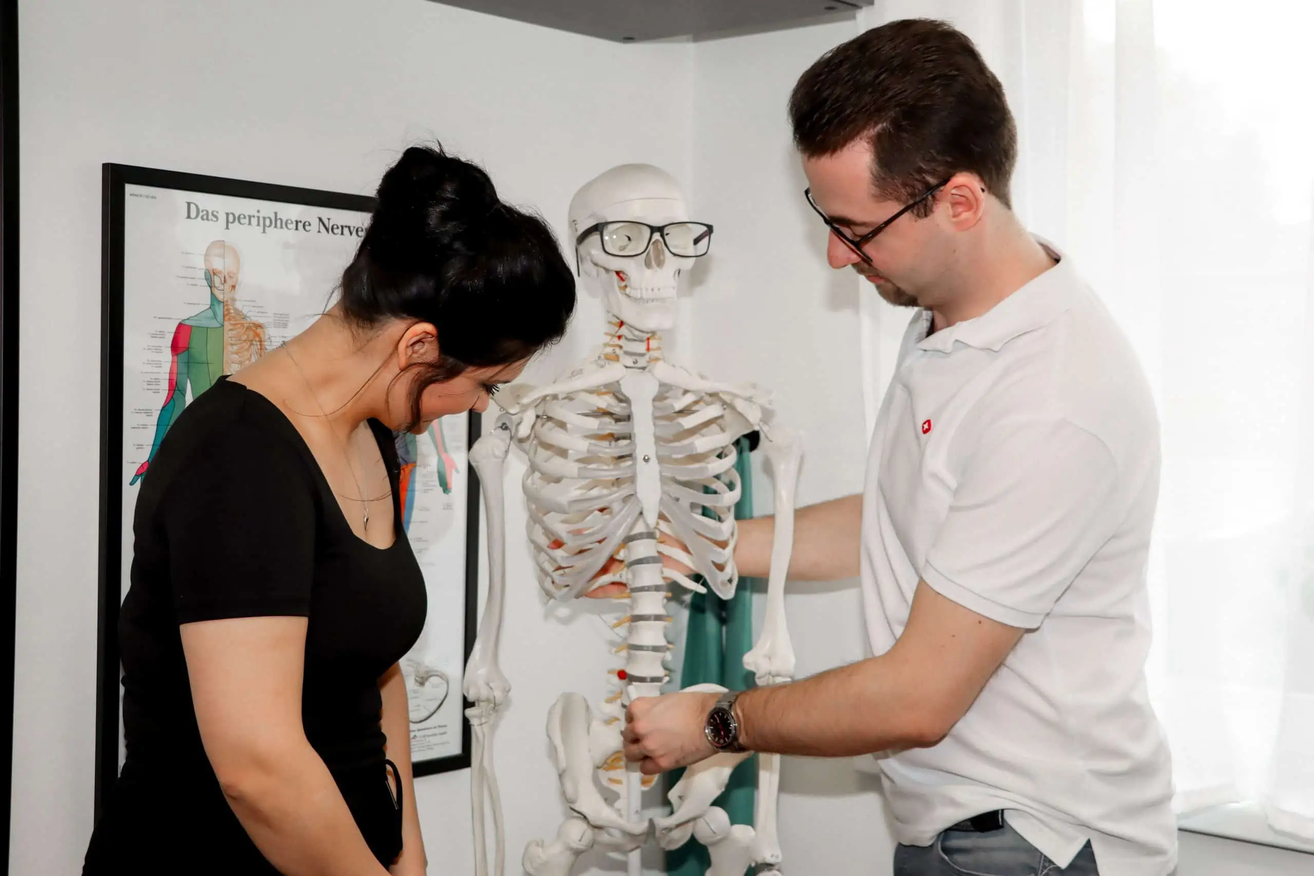 Zwei Personen untersuchen ein menschliches Skelettmodell, eine davon trägt eine Brille. Im Hintergrund ist ein Poster des Nervensystems zu sehen.
