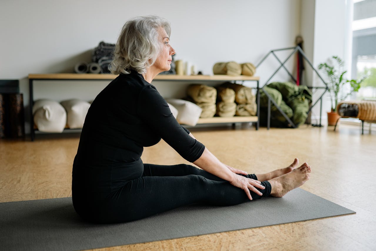 Eine ältere Frau in schwarzer Sportkleidung sitzt drinnen auf einer Yogamatte und macht leichte Übungen, während sie sich nach vorne streckt, um ihre Beine zu berühren, mit Yoga-Requisiten und Pflanzen im Hintergrund.