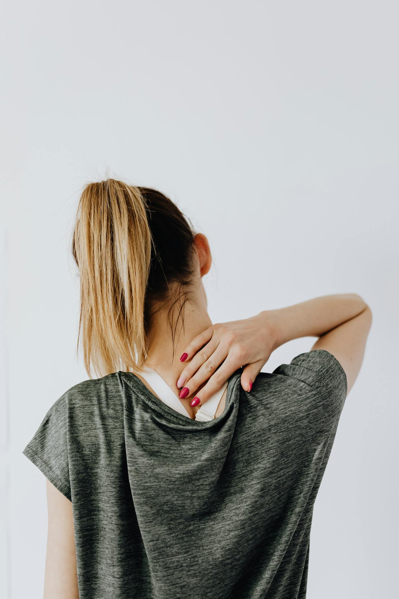 Eine Frau mit Pferdeschwanz, von hinten gesehen, berührt mit der linken Hand ihren Nacken, während sie ein graues T-Shirt trägt, und weist so auf mögliche Verspannungen in der Nacken- und Lendenwirbelsäule hin.