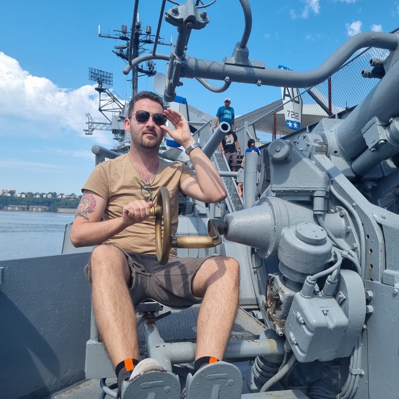 Ein Mann sitzt auf einem grauen Marinegeschütz, hält das Steuerrad und salutiert vor blauem Himmel und Wasser im Hintergrund.